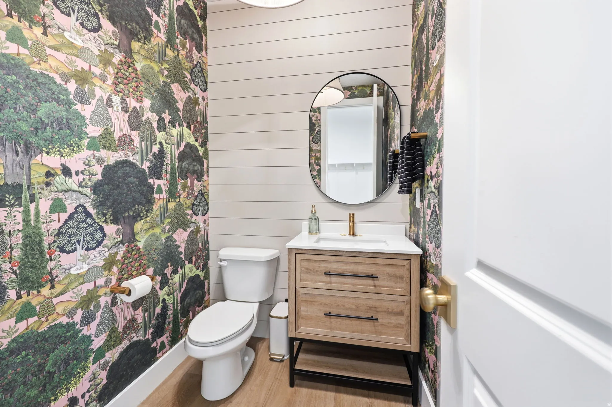 Powder room featuring vibrant botanical wallpaper on accent walls, white shiplap paneling, and a wood-finish vanity with two drawers and brass hardware
