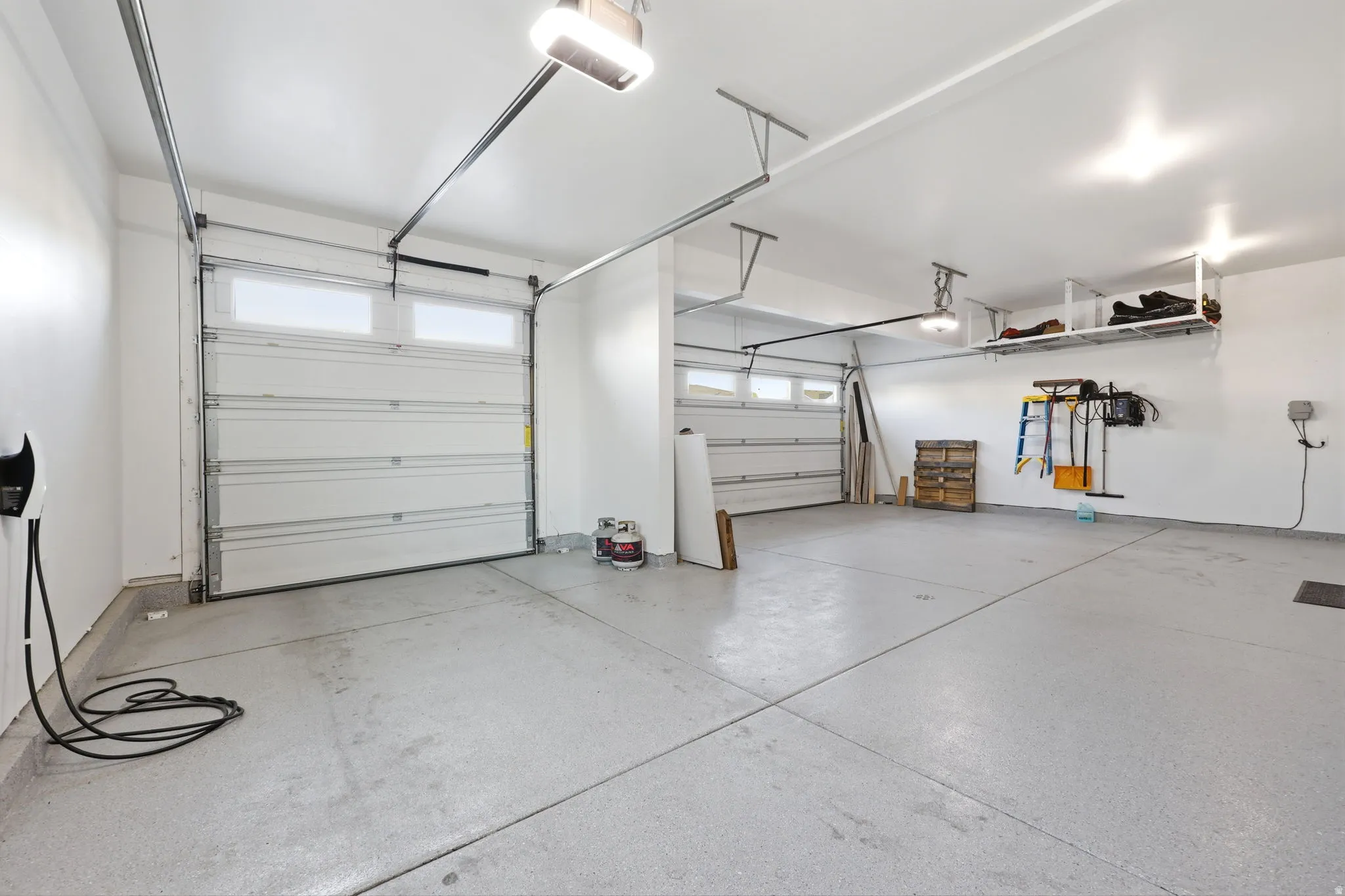 Spacious two-car garage featuring epoxy-coated concrete flooring, overhead storage systems, integrated LED lighting, and a side entry door with natural light from dual garage doors