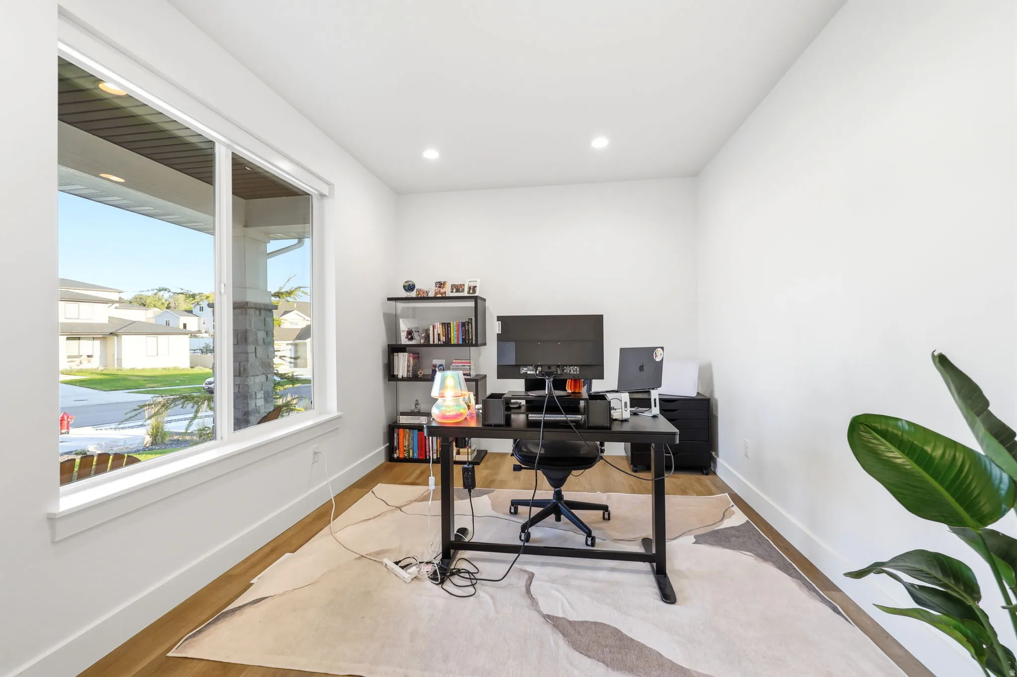 Dedicated home office featuring a large picture window with natural light, custom black desk with cable management, built-in shelving with storage, recessed ceiling lighting, wood-finish flooring, and a geometric area rug