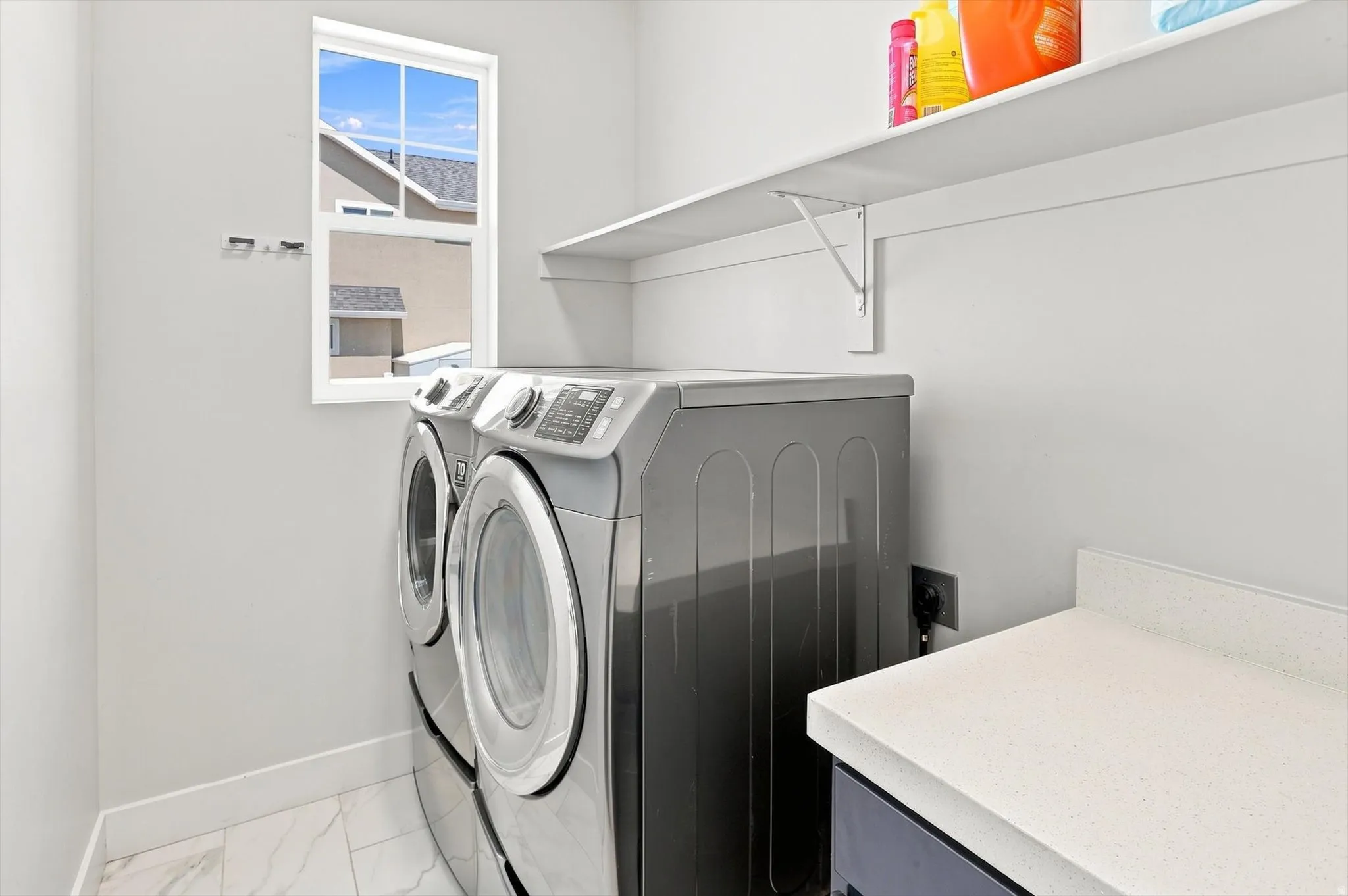 Modern laundry room featuring front-load washer and dryer with digital controls, white tile flooring, floating shelving with storage, and a utility sink with white countertop