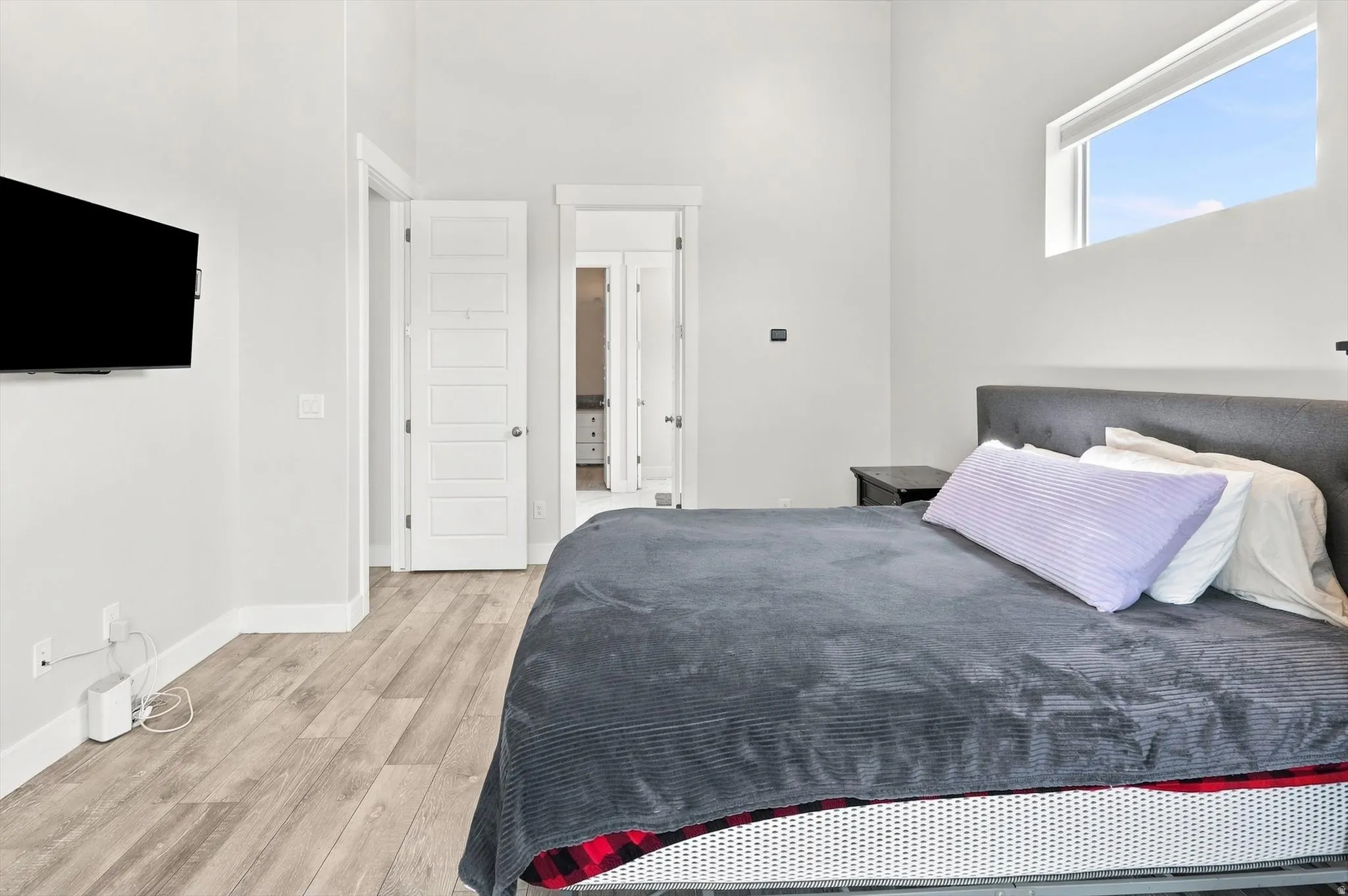 Modern primary bedroom featuring a wood-finish plank floor, contemporary gray upholstered headboard, large transom window with natural light, white panel doors with louvered detailing, and a wall-mounted flat-screen television
