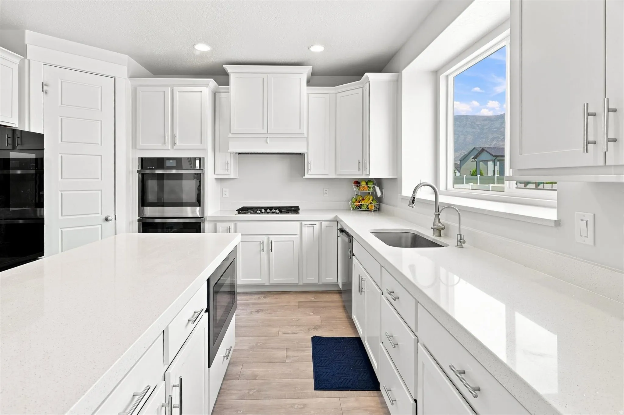 Contemporary kitchen featuring white shaker-style cabinetry, white quartz countertops, stainless steel appliances including a gas cooktop and double wall ovens, an undermount sink with chrome faucet, and wood-finish flooring throughout