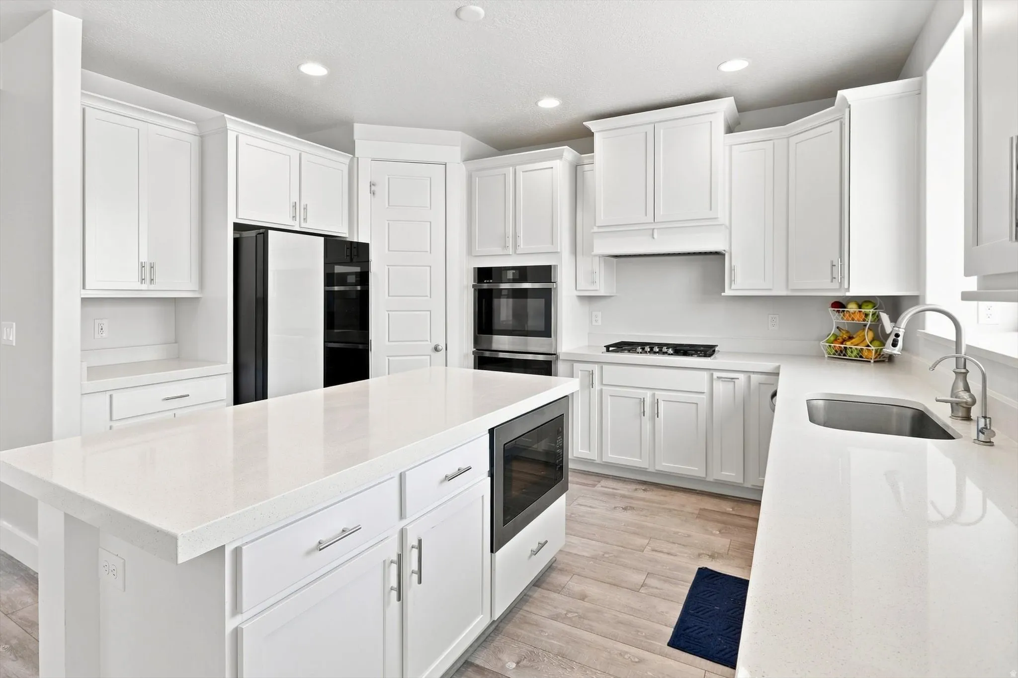 White cabinetry, light-toned wood-finish flooring, built-in double ovens, stainless steel sink, and a large central island