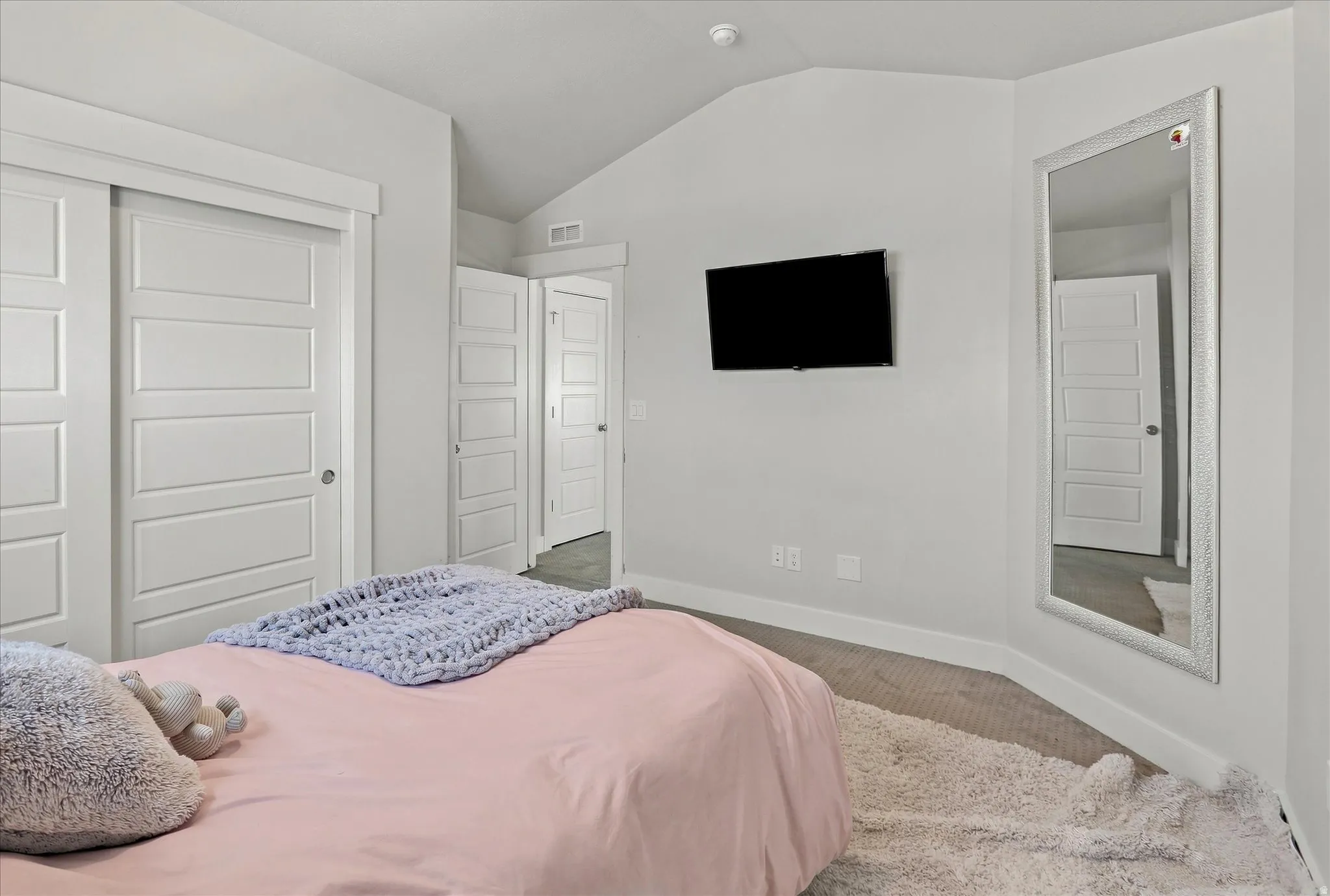 Bright primary bedroom featuring clean white finishes, recessed ceiling detail, wall-mounted flat-screen television, mirrored closet doors, and wood-finish flooring throughout