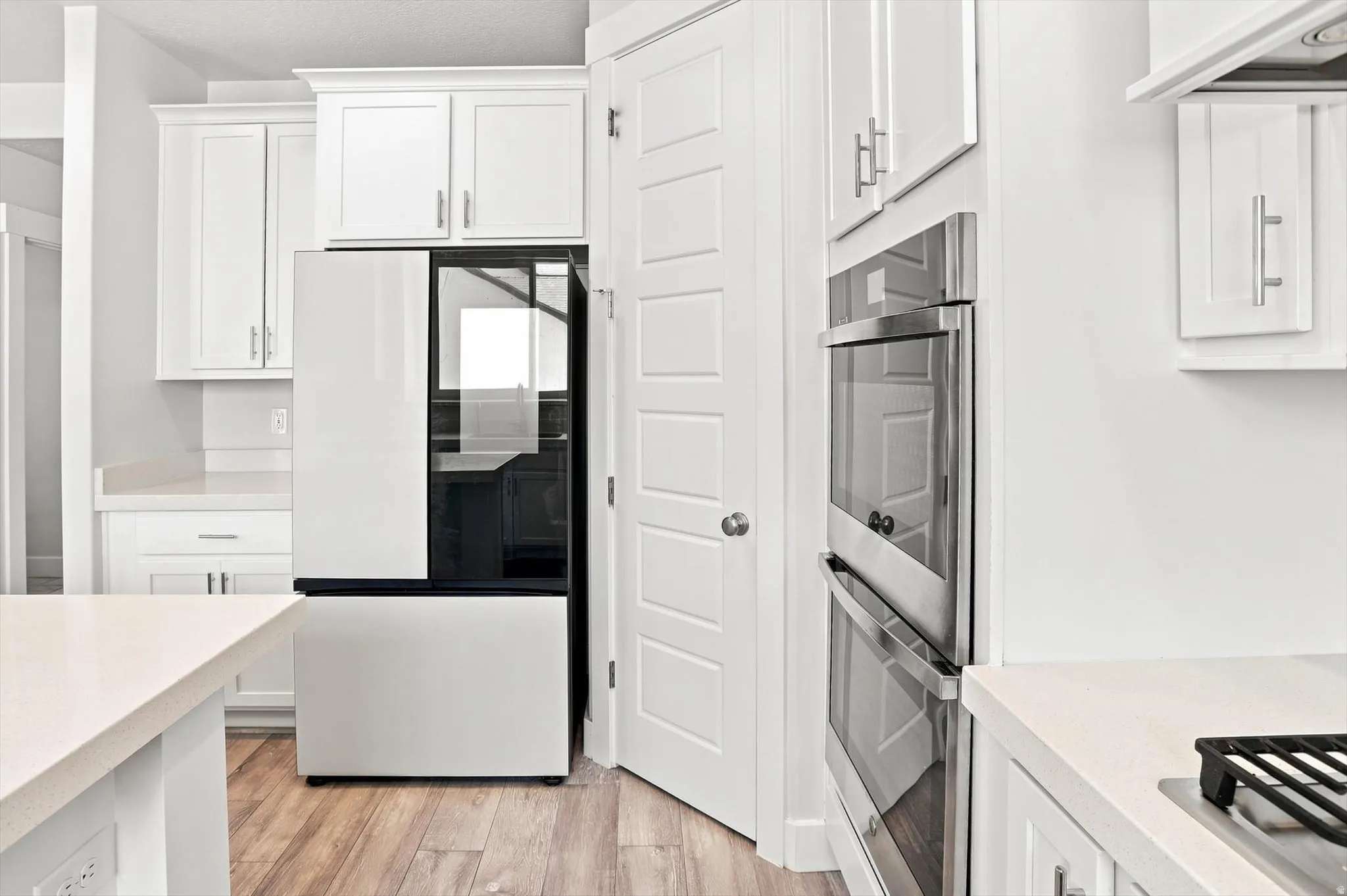 Modern kitchen featuring white cabinetry with stainless steel hardware, wood-finish flooring throughout, dual wall-mounted stainless steel ovens, and a sleek black refrigerator with glass door panel