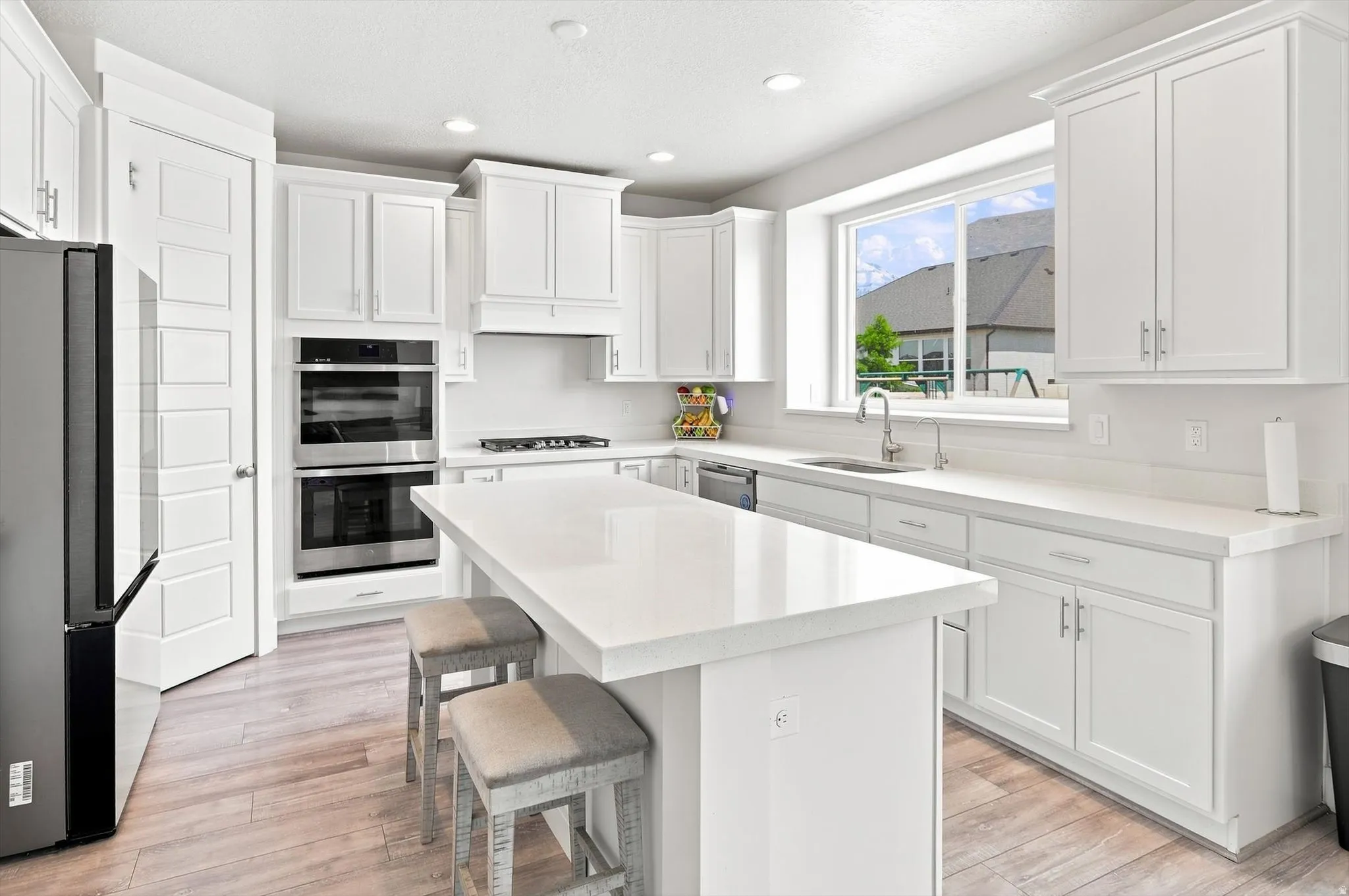 Contemporary kitchen featuring white shaker-style cabinetry, a large quartz-topped center island with seating for two, stainless steel appliances including dual wall ovens and a gas cooktop, subway tile backsplash, and abundant natural light from expansiv