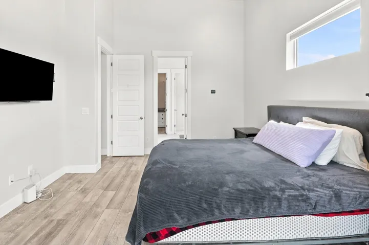 Modern primary bedroom featuring a wood-finish plank floor, contemporary gray upholstered headboard, large transom window with natural light, white panel doors with louvered detailing, and a wall-mounted flat-screen television