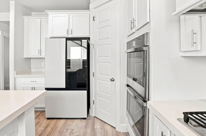 Modern kitchen featuring white cabinetry with stainless steel hardware, wood-finish flooring throughout, dual wall-mounted stainless steel ovens, and a sleek black refrigerator with glass door panel