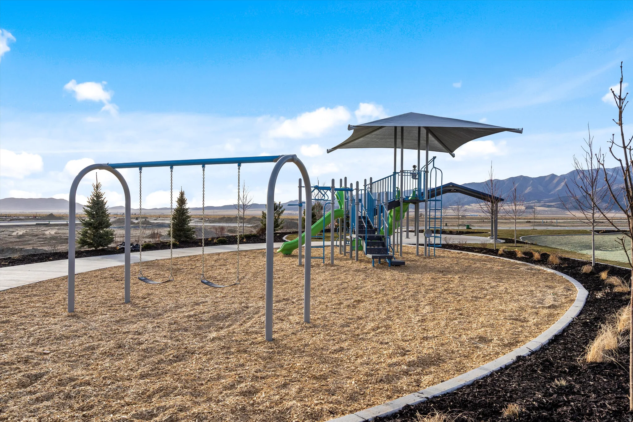 Playground area featuring a swing set, multi-level play structure with slides, and a shaded seating area, surrounded by woodchip surfacing and a paved pathway