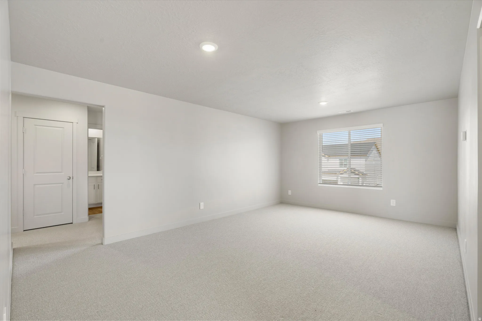 **DISCLAIMER** Interior package may vary. Bright, generously proportioned bedroom featuring neutral carpet flooring, recessed ceiling lighting, a large window with horizontal blinds providing natural light, and a white six-panel closet door with adjacent
