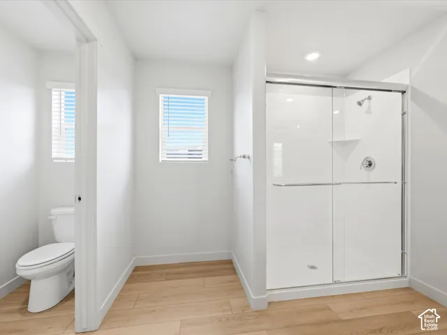 Walk-in shower with a glass enclosure, wood-finish flooring, two windows with blinds, and a standard-height toilet