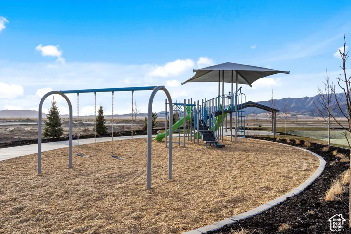 Playground area featuring a swing set, multi-level play structure with slides, and a shaded seating area, surrounded by woodchip surfacing and a paved pathway
