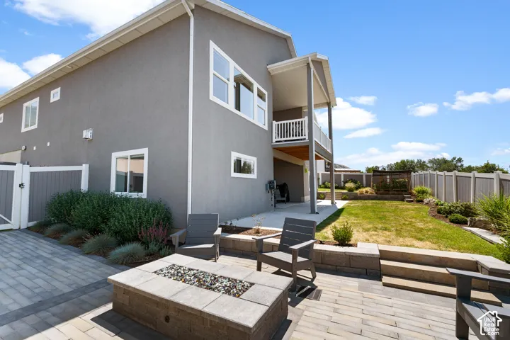 Expansive backyard featuring a paver patio with a built-in fire pit, a tiered lawn area with landscaping, a privacy fence, and a covered patio area below a second-story balcony