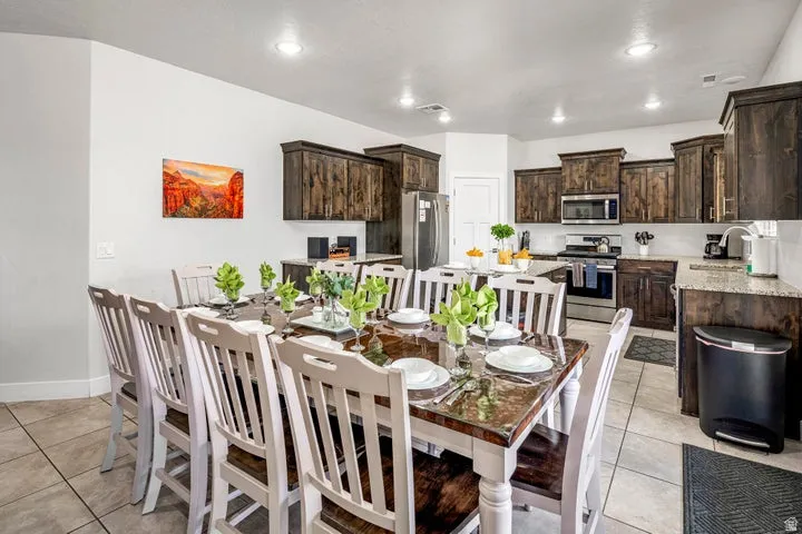 Open-concept dining and kitchen area featuring a wood-finish dining table with seating for eight, tile flooring throughout, dark wood cabinetry with stainless steel appliances, white subway tile backsplash, and recessed ceiling lighting