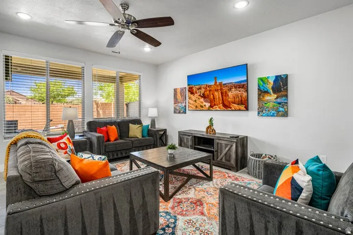Bright living room featuring multiple seating areas with gray upholstered furnishings, a ceiling fan with integrated lighting, and large windows with plantation shutters providing natural light