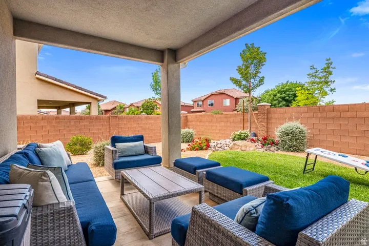 Covered patio with concrete flooring and structural columns opening to a spacious backyard featuring mature landscaping, manicured lawn, brick perimeter fencing, and woven outdoor seating arrangements with cushioned elements