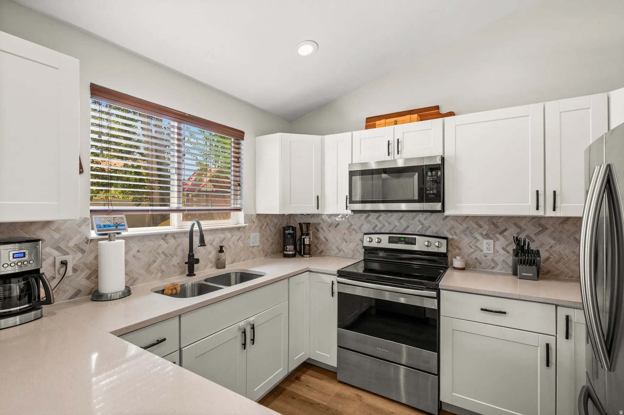 Modern kitchen featuring white shaker cabinetry, stainless steel appliances, light-toned countertops, a herringbone tile backsplash, and wood-finish flooring