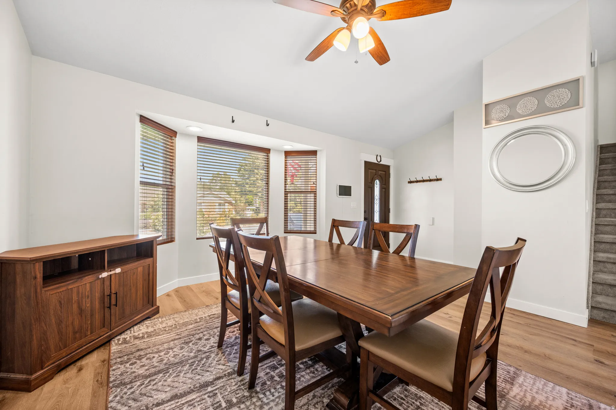 Formal dining area featuring a bay window with blinds, wood-finish flooring, a ceiling fan, and staircase access