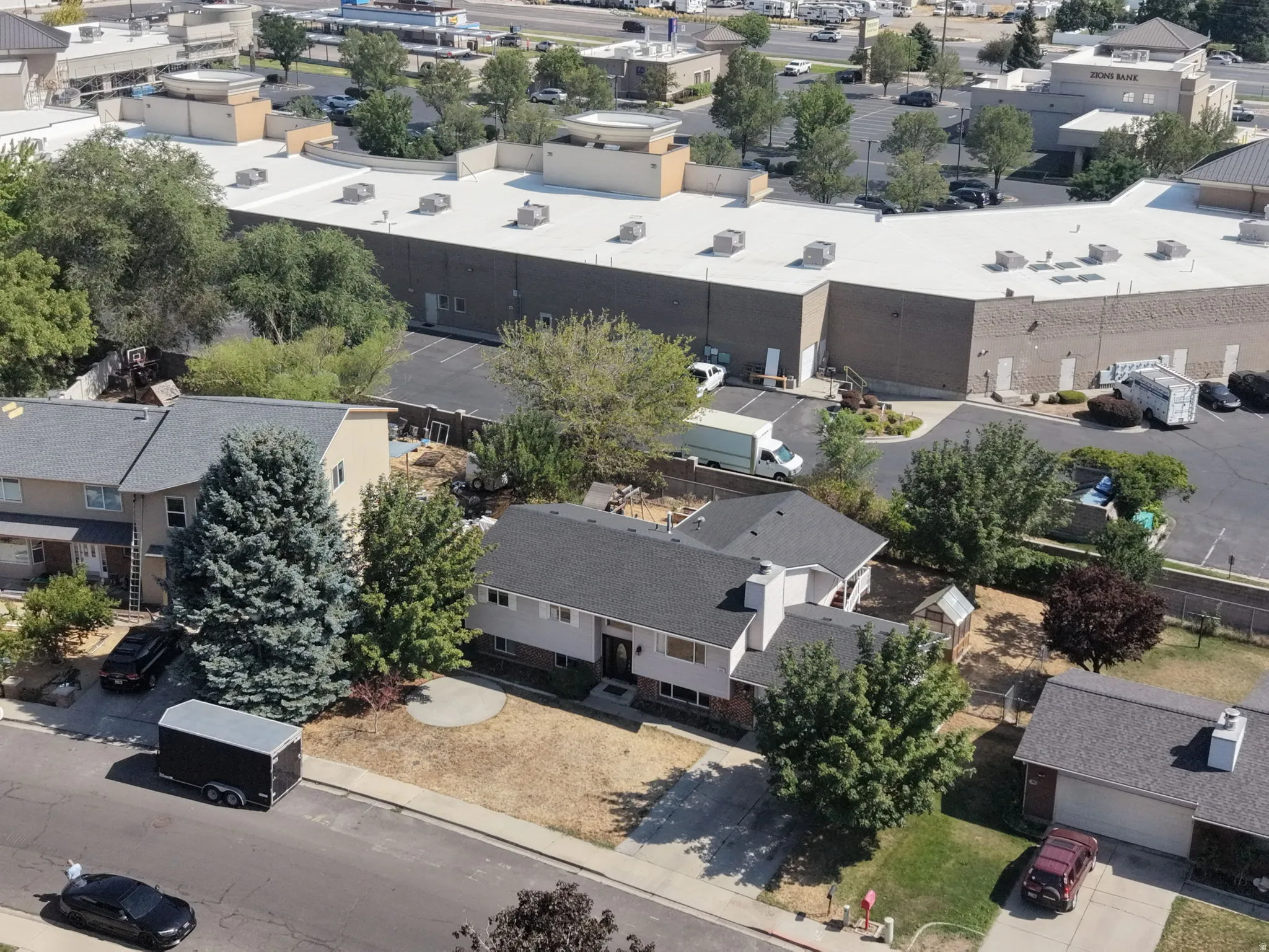 Aerial view of a residential property featuring a single-story residence with a flat roof, surrounded by mature landscaping including evergreen and deciduous trees