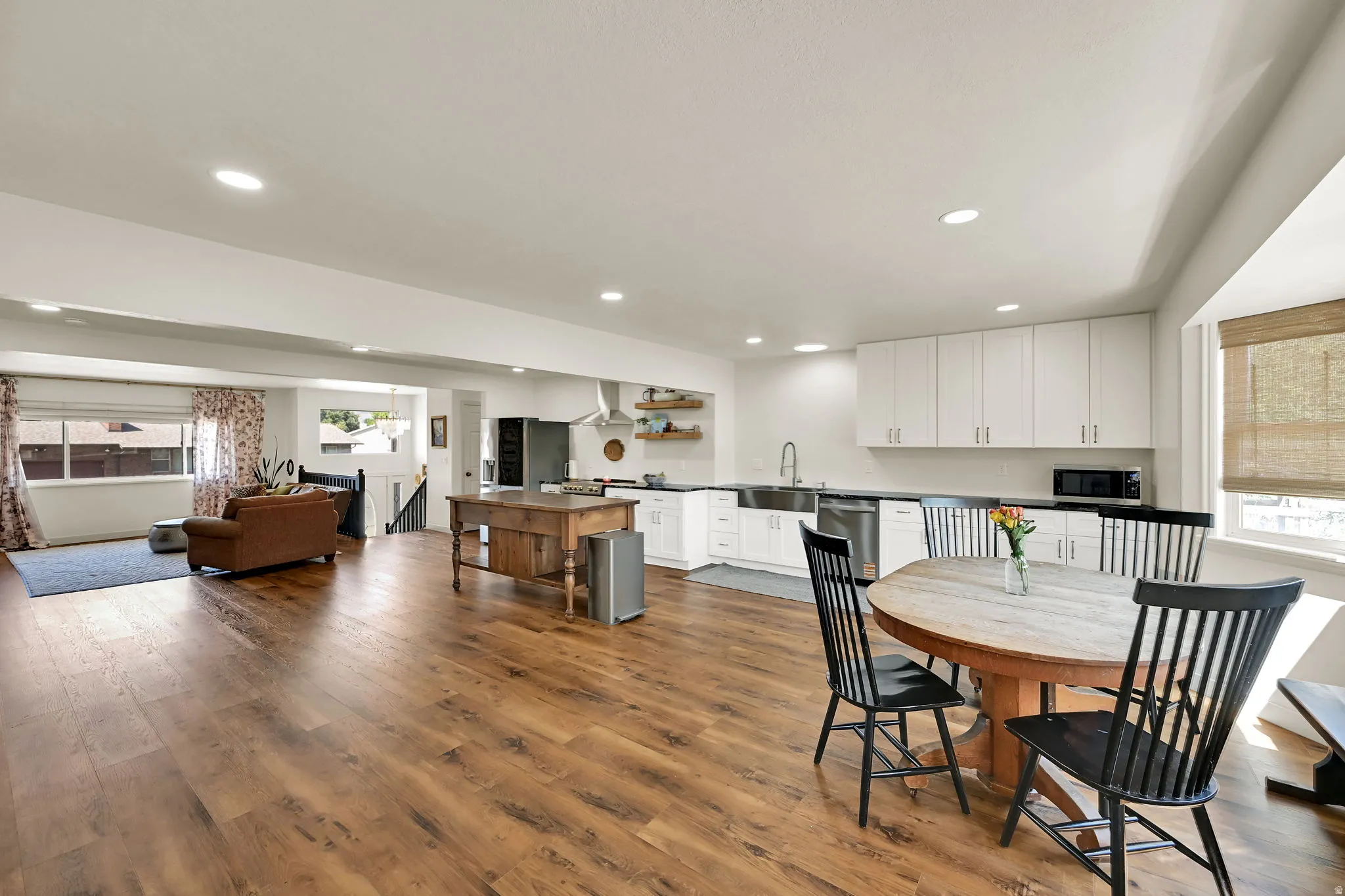 Open-concept living space featuring warm wood-finish flooring throughout, white cabinetry with integrated appliances, a central island with seating, recessed ceiling lighting, and abundant natural light from multiple windows