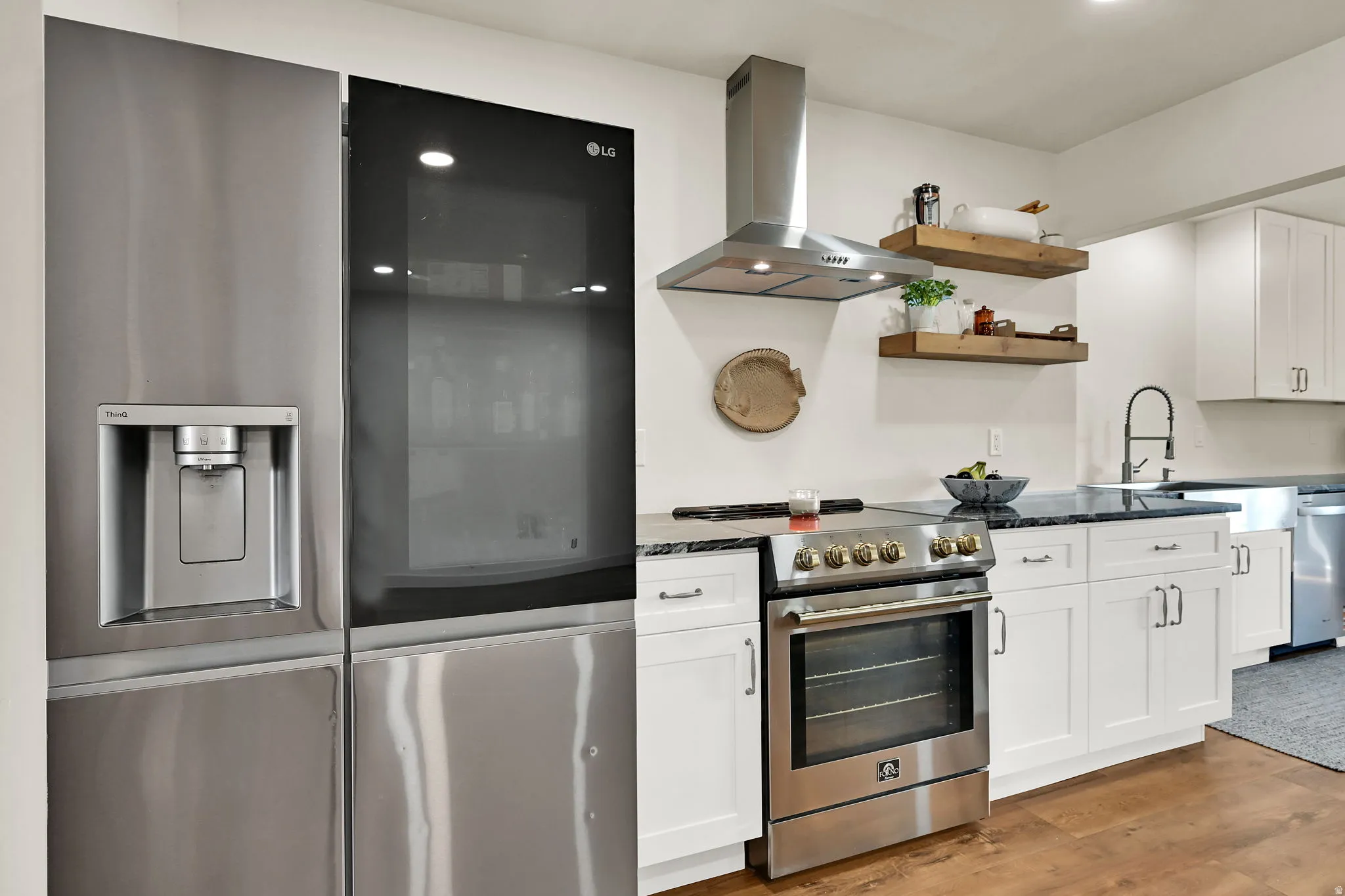 Stainless steel refrigerator with integrated ice and water dispenser, stainless steel range with overhead vent hood, white shaker-style cabinetry, dark stone countertops, and wood-finish flooring