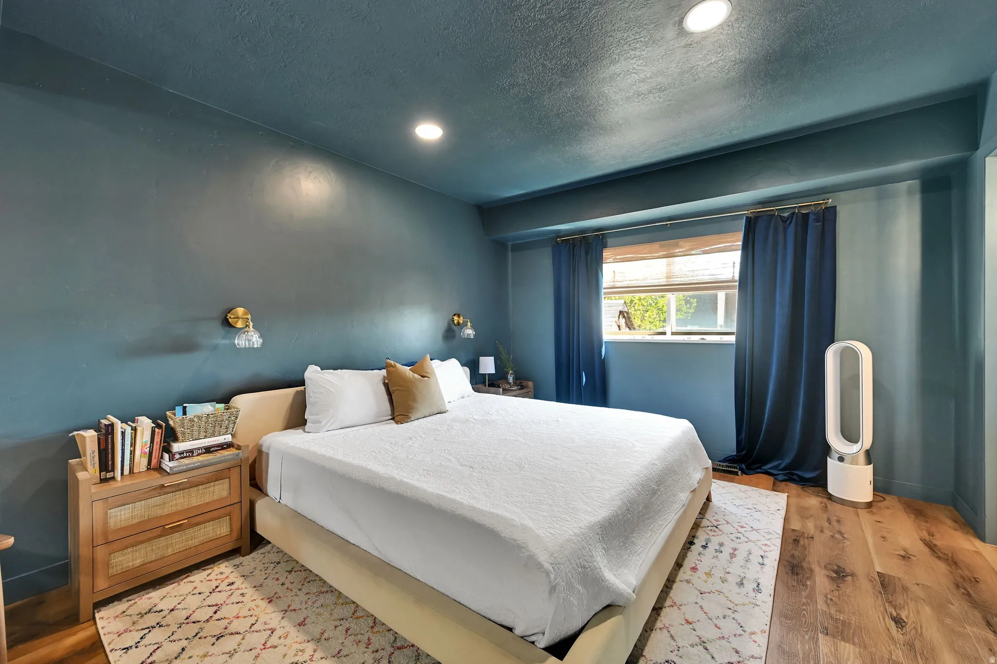 Serene primary bedroom showcasing soft blue-gray wall finishes, recessed ceiling lighting, wood-finish flooring, and geometric patterned area rug