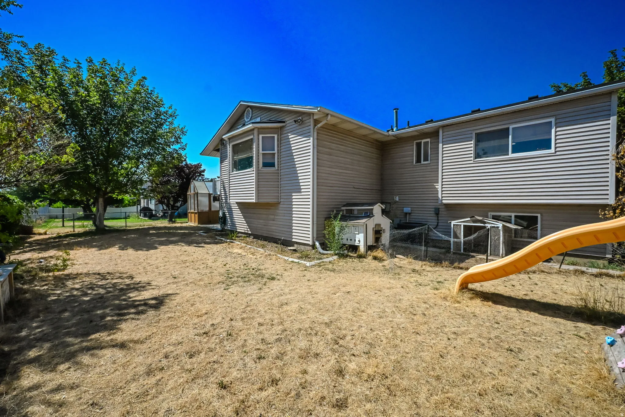 Spacious backyard with mature shade trees and level sandy grounds, complemented by the home's tan vinyl siding exterior and multi-section roofline