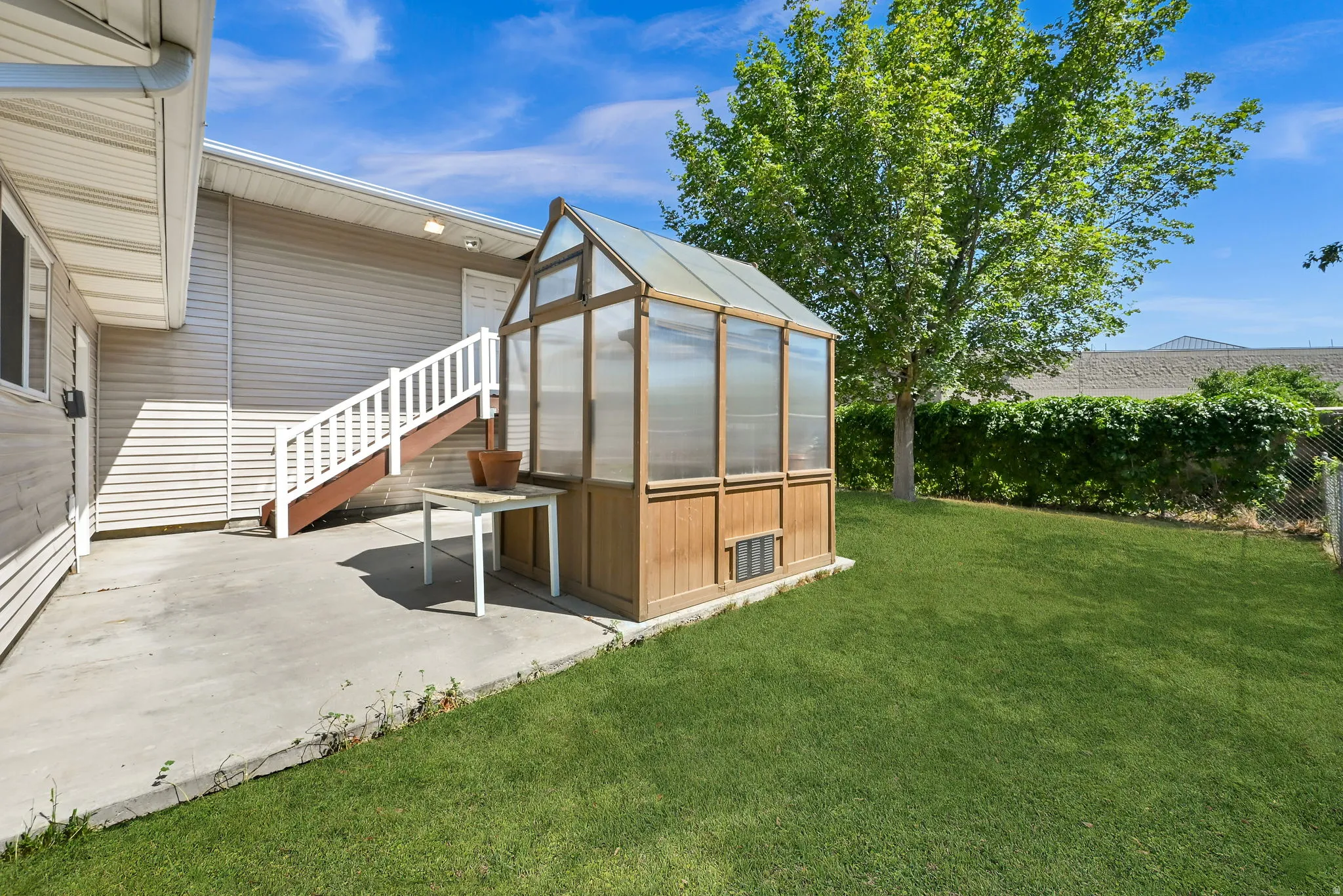 Spacious backyard oasis featuring a glass-paneled greenhouse structure with wood-finish base, expansive manicured lawn, mature shade trees, and concrete patio adjoining the home
