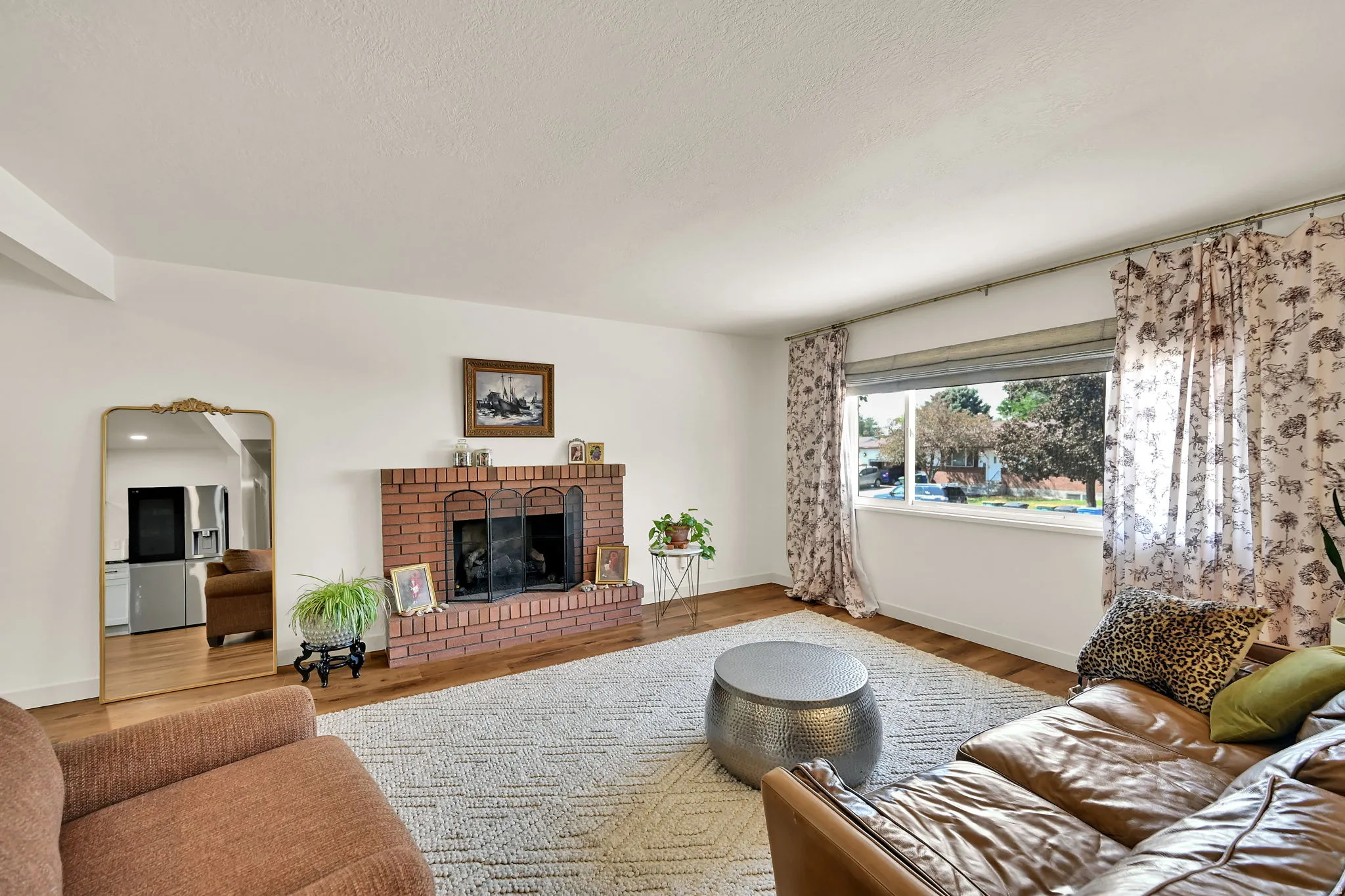 Brick fireplace with a metal screen, wood-finish flooring, and a large window with roller shades
