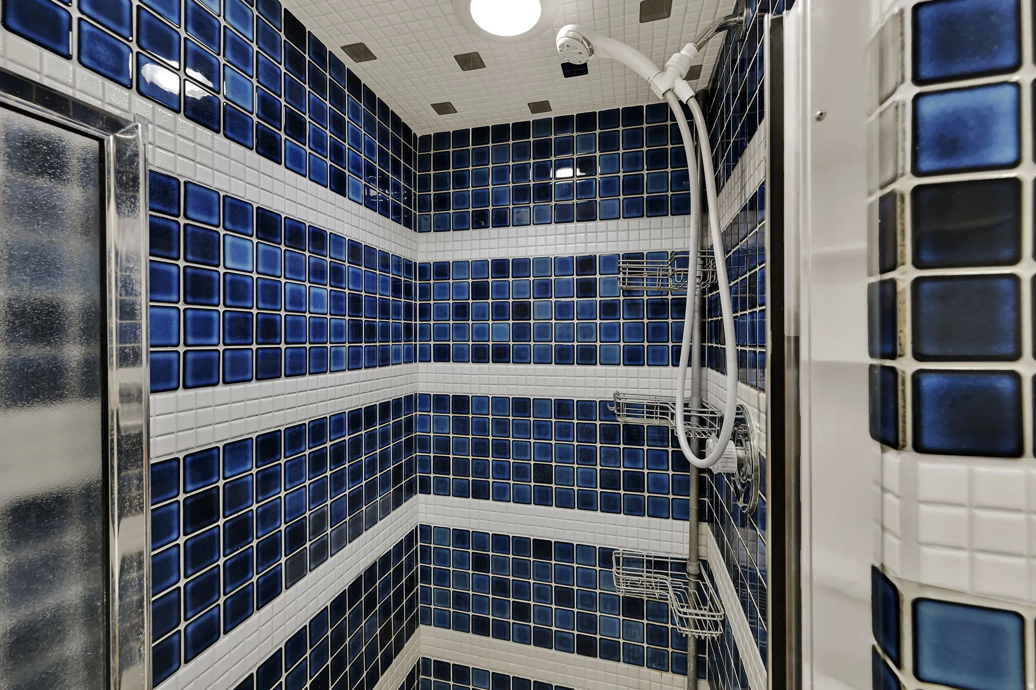 Modern shower enclosure featuring deep navy blue tile walls with contrasting white tile accents, chrome showerhead and fixtures, and recessed ceiling lighting for a spa-like bathing experience