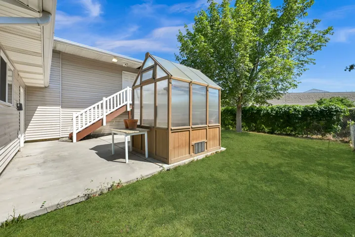 Spacious backyard oasis featuring a glass-paneled greenhouse structure with wood-finish base, expansive manicured lawn, mature shade trees, and concrete patio adjoining the home