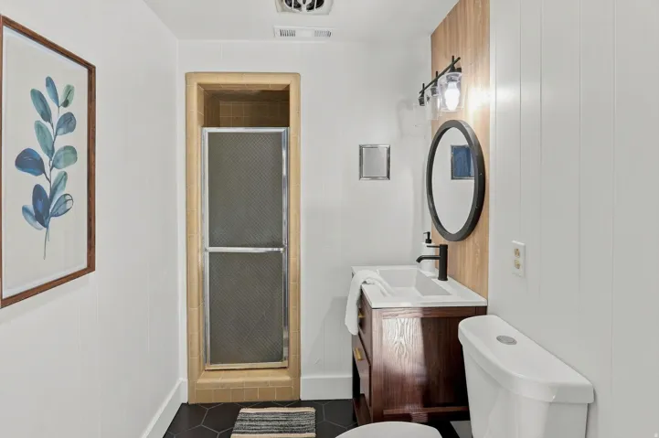 Compact bathroom featuring a wood-finish vanity with integrated sink, circular mirror with matte black frame, white ceramic toilet, and dark tile flooring