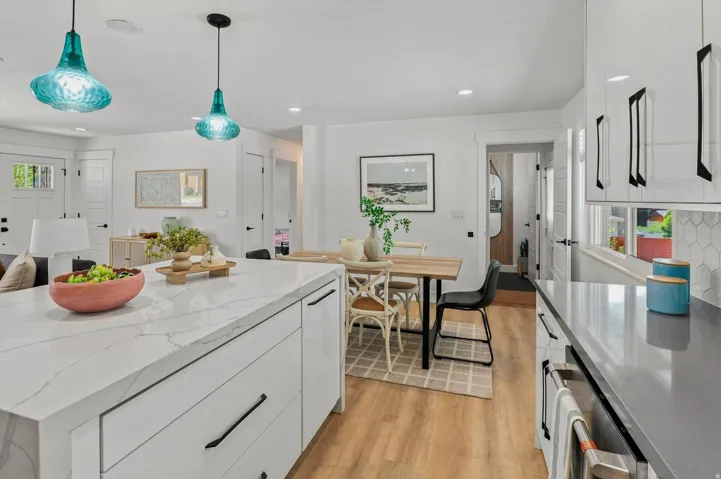 Kitchen featuring a large waterfall island with a light-toned countertop, white cabinetry with black hardware, wood-finish flooring, and two pendant light fixtures