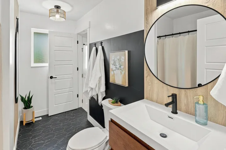 Black hexagon tile flooring, a dark accent wall, a wood-finish vanity with a rectangular basin sink, a circular black-framed mirror, and a frosted window