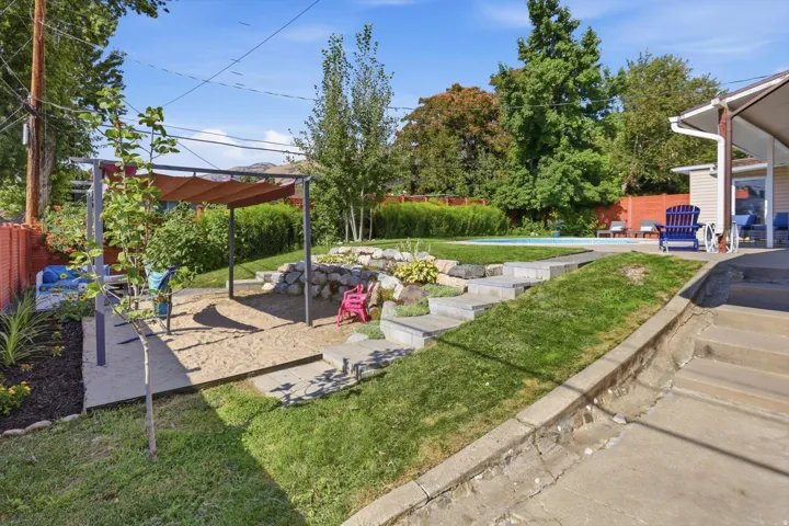 Multi-tiered backyard featuring a swimming pool, stone retaining walls, concrete steps, and a pergola-covered sand area