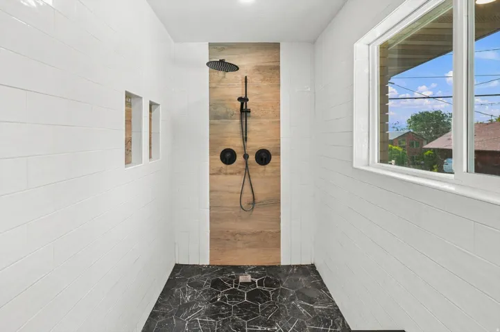 Contemporary primary bathroom featuring a wood-finish accent wall, white subway tile surround, polished dark stone flooring with geometric patterning, dual matte black showerheads with handheld wand, and large-format window with natural light and exterior