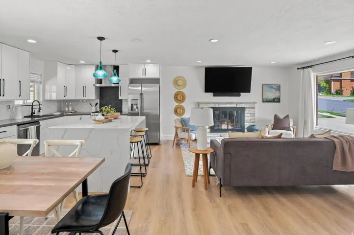 Open-concept living space featuring white cabinetry and stainless steel appliances in the kitchen, wood-finish flooring throughout, pendant lighting, a wall-mounted flat-screen television, and a fireplace with built-in surround in the living area