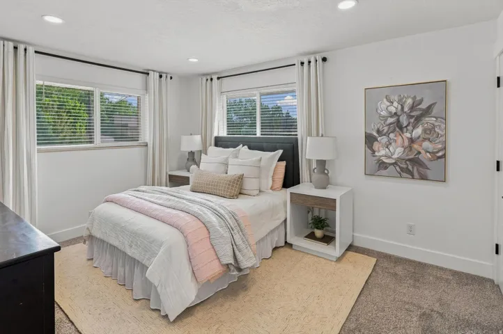 Bright secondary bedroom featuring multiple windows with natural light, neutral wall finishes, wood-finish flooring, and a built-in nightstand with integrated lighting