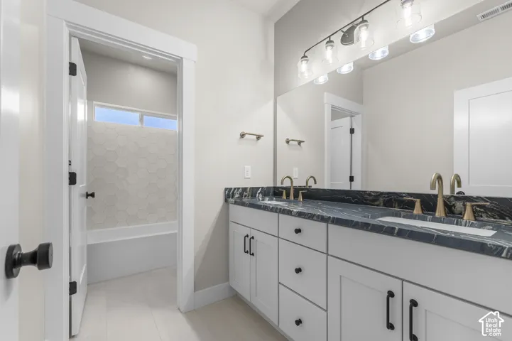 Double-sink vanity featuring a stone countertop, white shaker-style cabinetry, matte black hardware, and metallic faucets, complemented by a large wall-mounted mirror and hexagonal tile bathtub surround