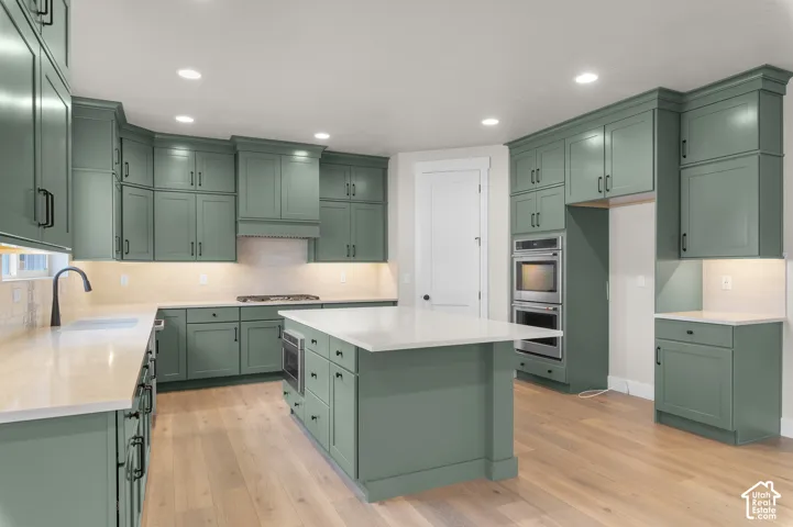 Kitchen featuring sage green shaker-style cabinetry, a central island with a solid-surface countertop, recessed ceiling lighting, double wall ovens, and wood-finish flooring