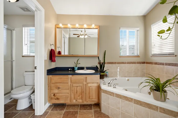 Corner jetted tub with tiled surround, wood-finish vanity with countertop, medicine cabinet, and tiled flooring