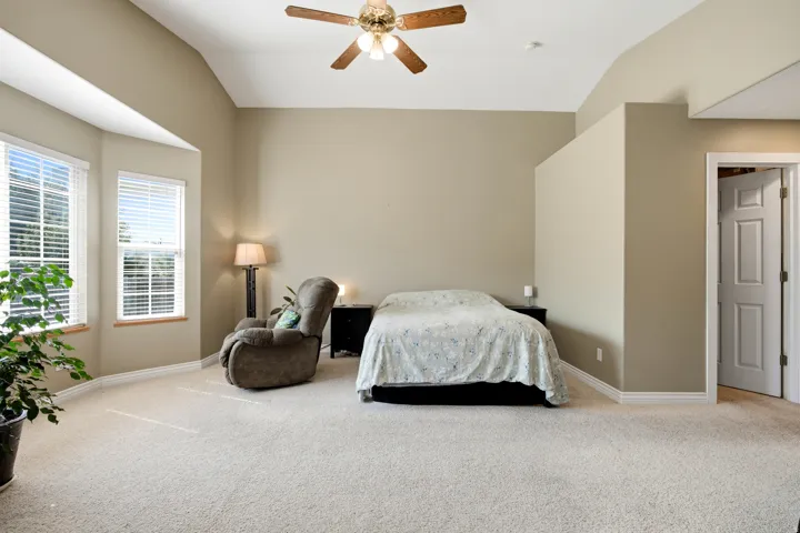 Vaulted ceiling, two large windows with blinds, wood-finish ceiling fan, neutral carpet flooring, and a paneled interior door