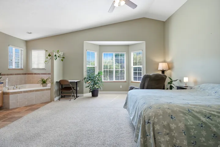 Vaulted ceilings with a ceiling fan, a bay window with blinds, a built-in tiled jetted tub, and carpet flooring