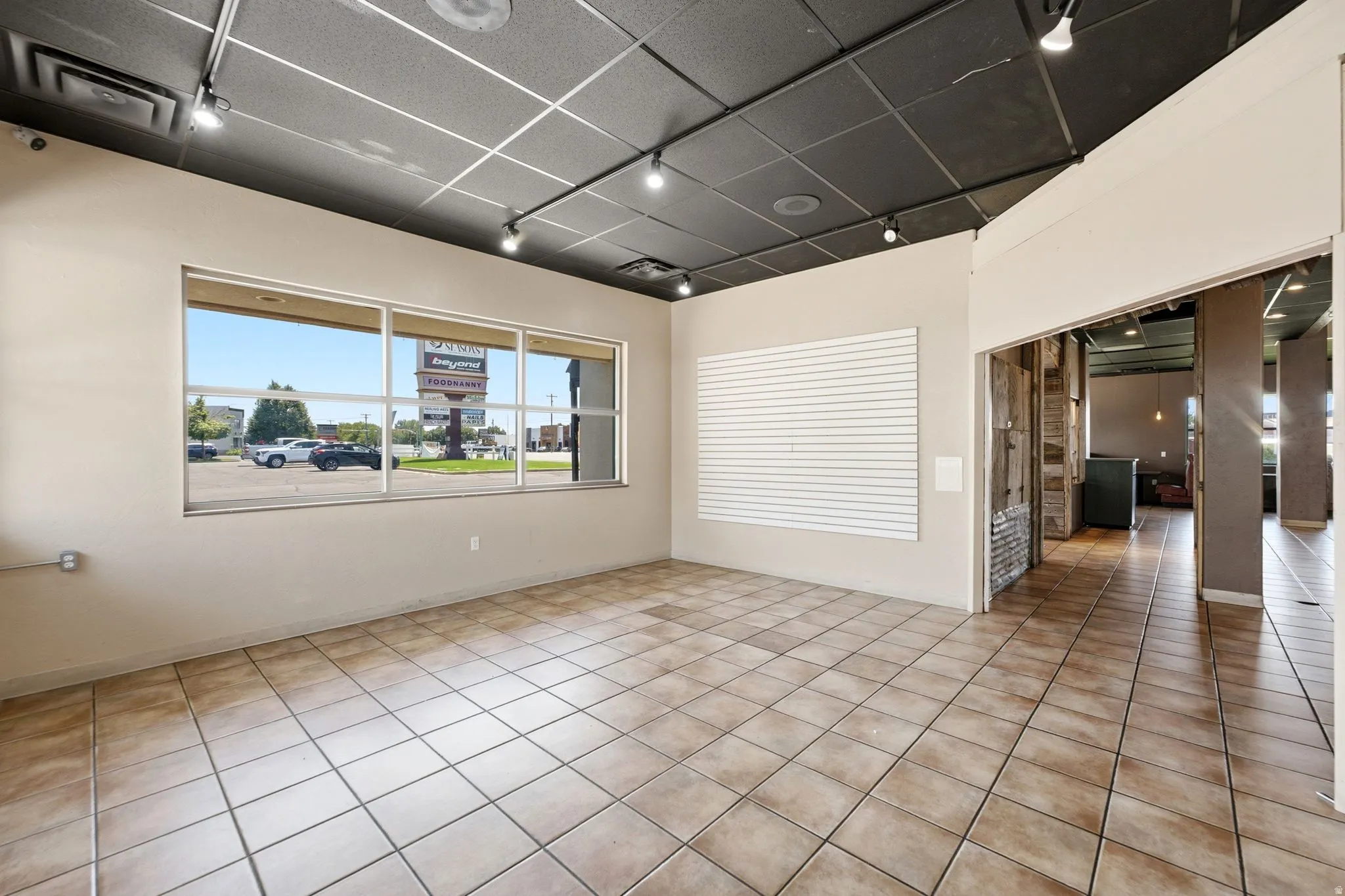 Bright corner retail or office space featuring expansive front windows with natural light, tile flooring throughout, recessed and track lighting in suspended ceiling, and direct access to adjacent interior spaces