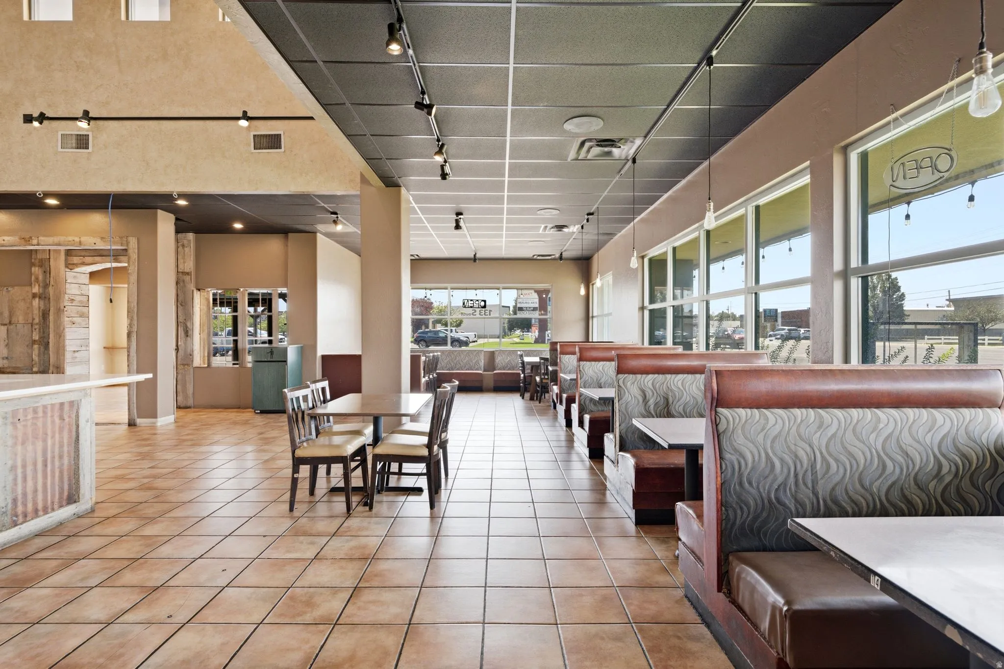 Spacious commercial or hospitality interior featuring expansive tile flooring, track lighting throughout a coffered ceiling, and floor-to-ceiling windows with natural light