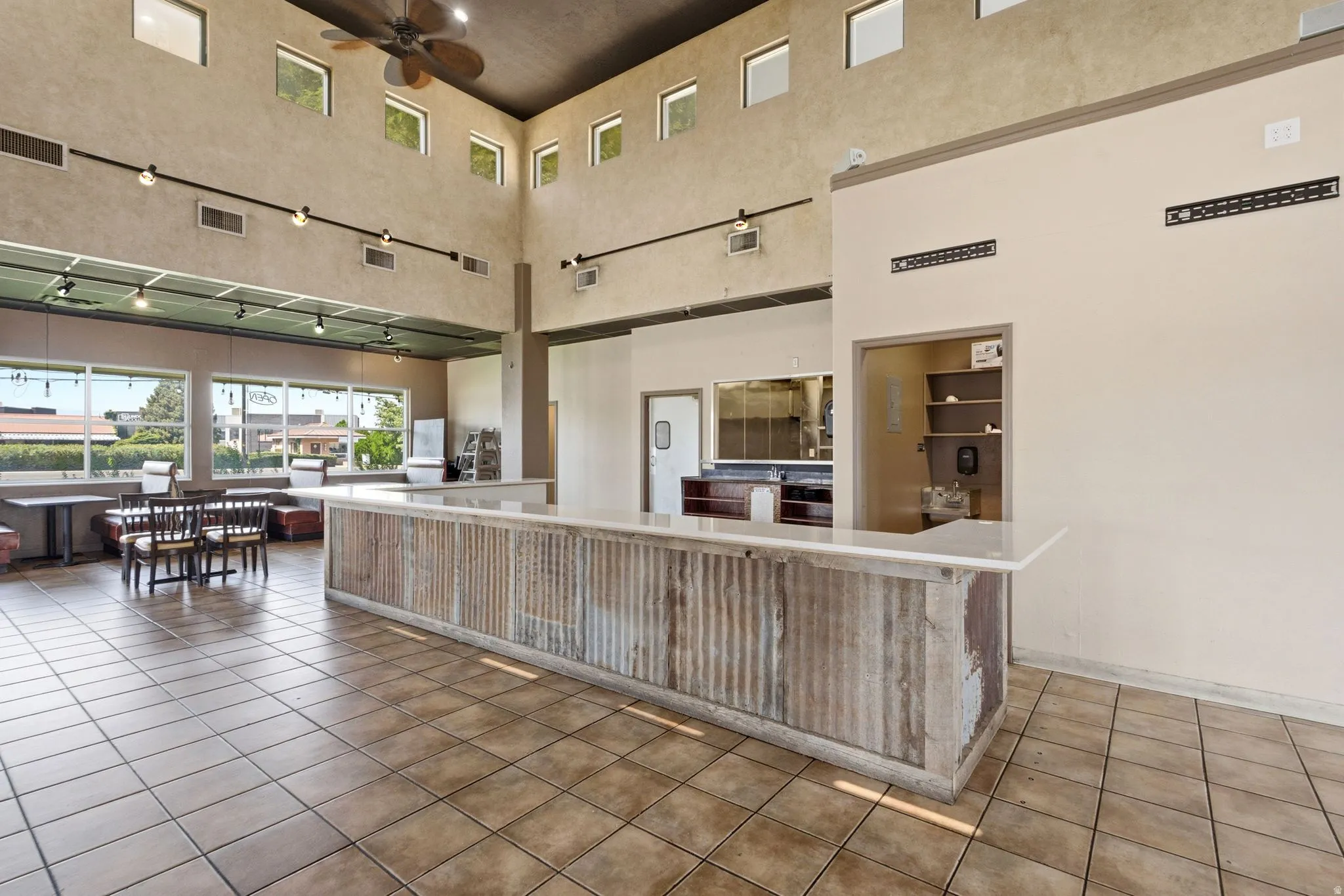 Spacious commercial or hospitality interior featuring a prominent wood-finish service counter with corrugated metal paneling, expansive tile flooring, high ceilings with clerestory windows and track lighting, and multiple service windows with wood-frame o