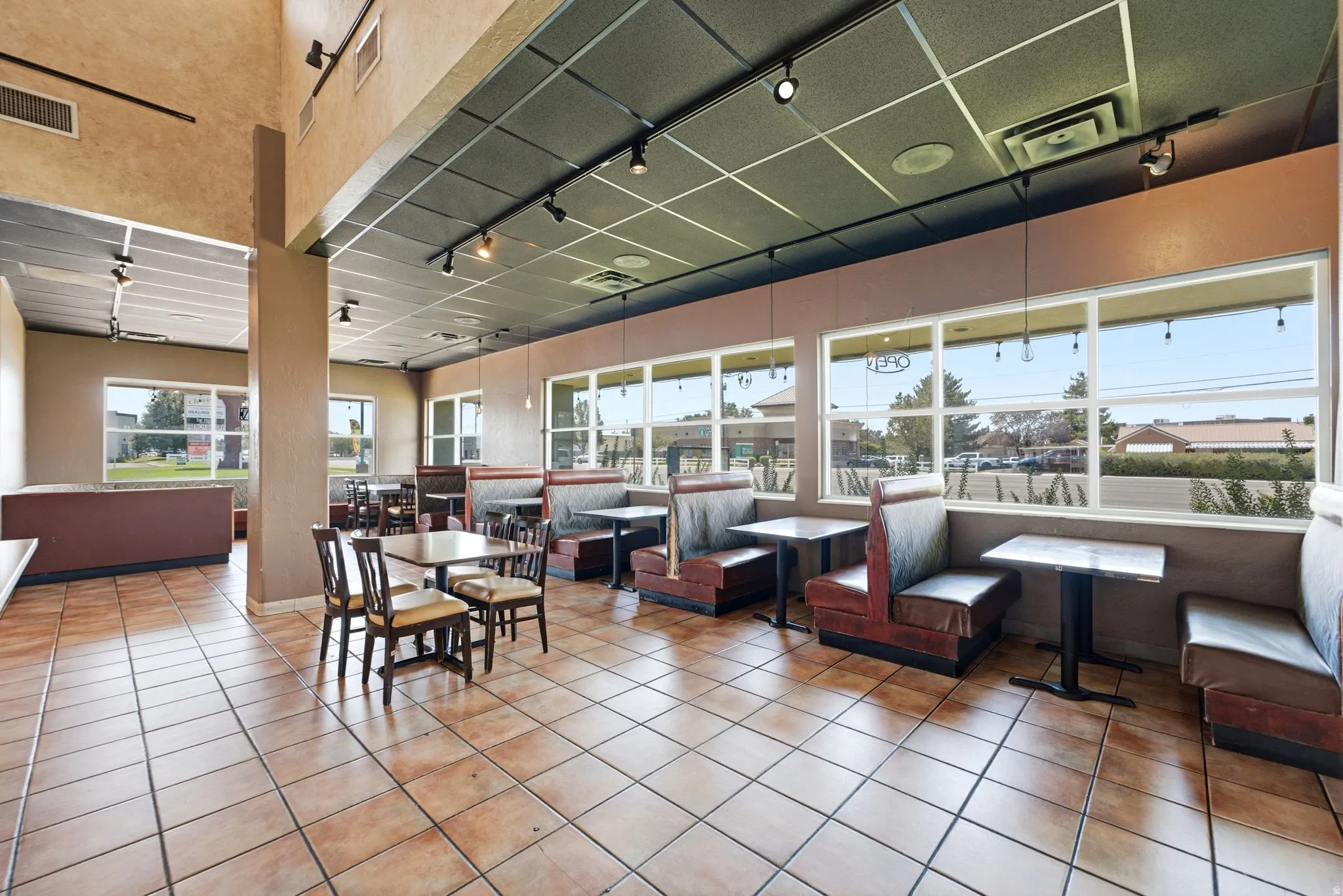 Bright, open-concept dining and lounge area featuring terracotta tile flooring, expansive windows with natural light, suspended ceiling with integrated lighting, and built-in upholstered banquette seating