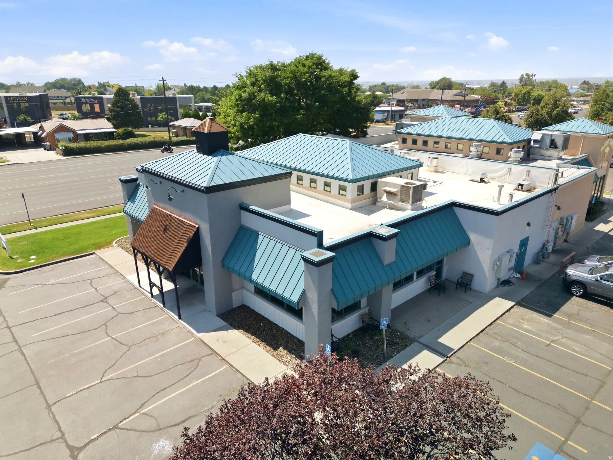 Modern commercial or mixed-use building featuring a distinctive angular roofline with standing-seam metal cladding in blue and copper tones, clean white exterior walls, and multiple roof-mounted mechanical units