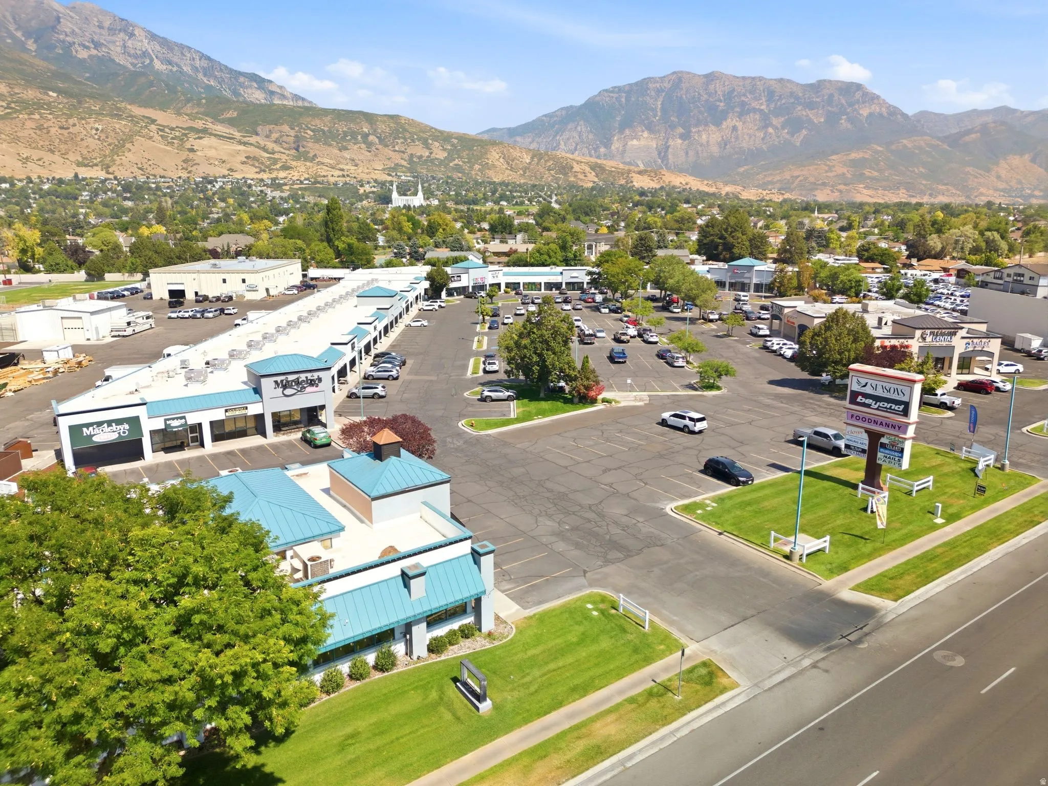 Commercial retail and office complex featuring multiple storefronts with modern architectural design, expansive asphalt parking lot with organized spaces, mature landscaping throughout the property, and striking mountain backdrop framing the entire develo