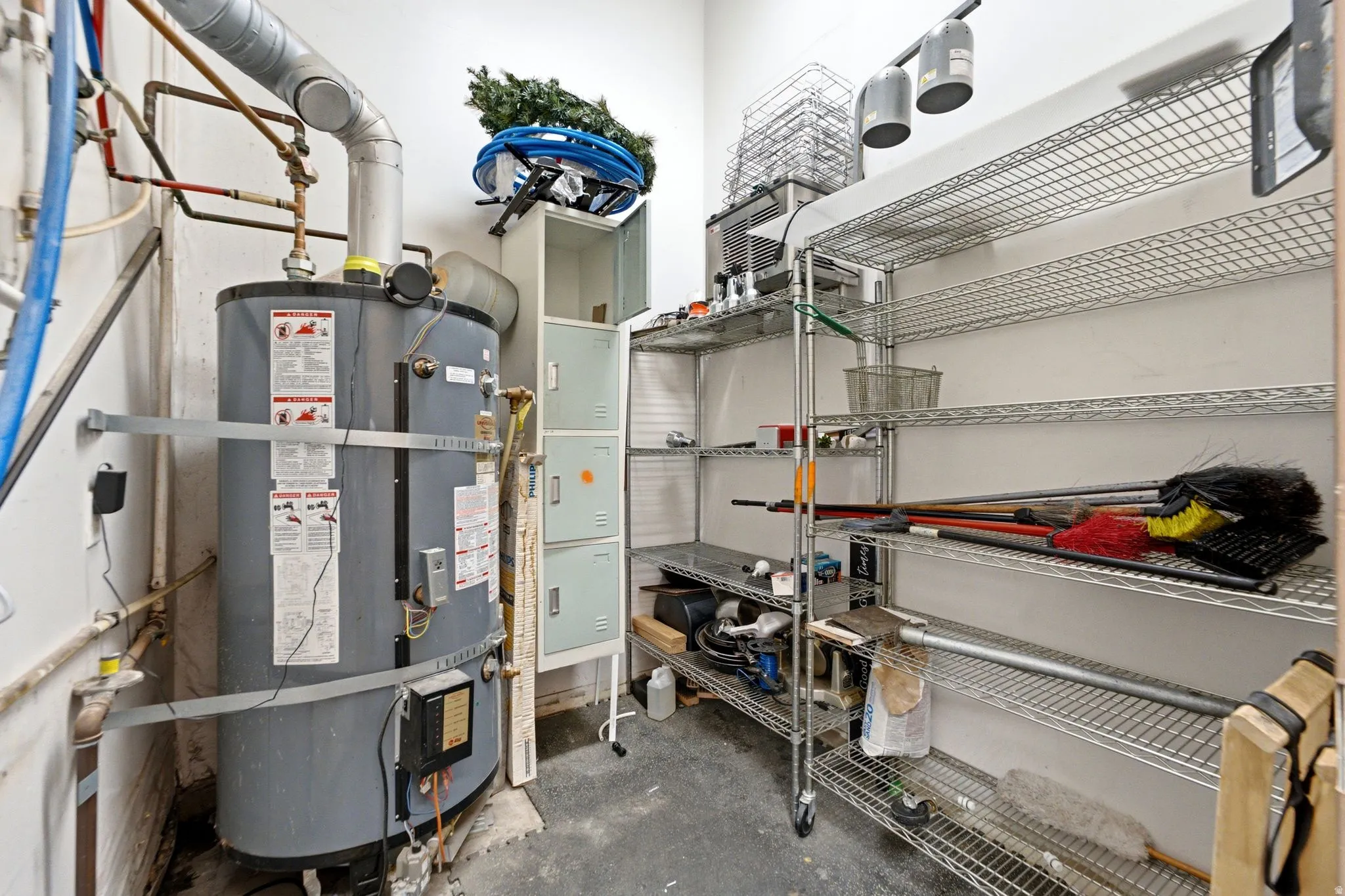 Utility room/dry storage featuring a high-capacity water heater, metal shelving with organized storage capacity, and overhead piping infrastructure